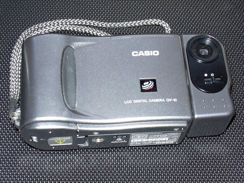 La cámara Casio QV-10 de 1995, gris, con el objetivo montado en un cuerpo giratorio a la derecha y el rótulo LCD DIGITAL CAMERA QV-10 impreso en el frontal.